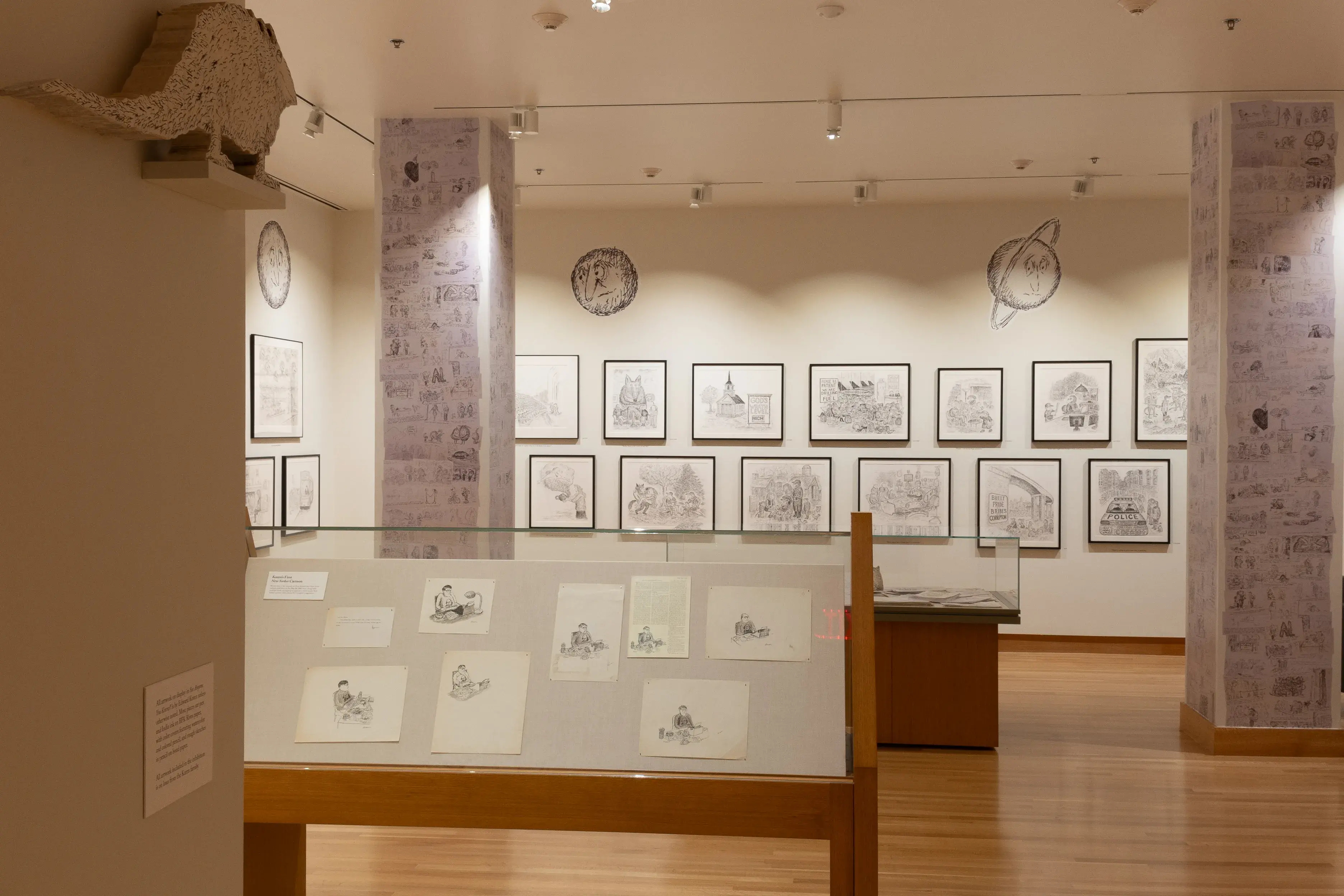 Installation image of a gallery exhibition. There are framed drawings on the wall and more drawings in a glass case in front. There are also drawings adhered to two square columns.