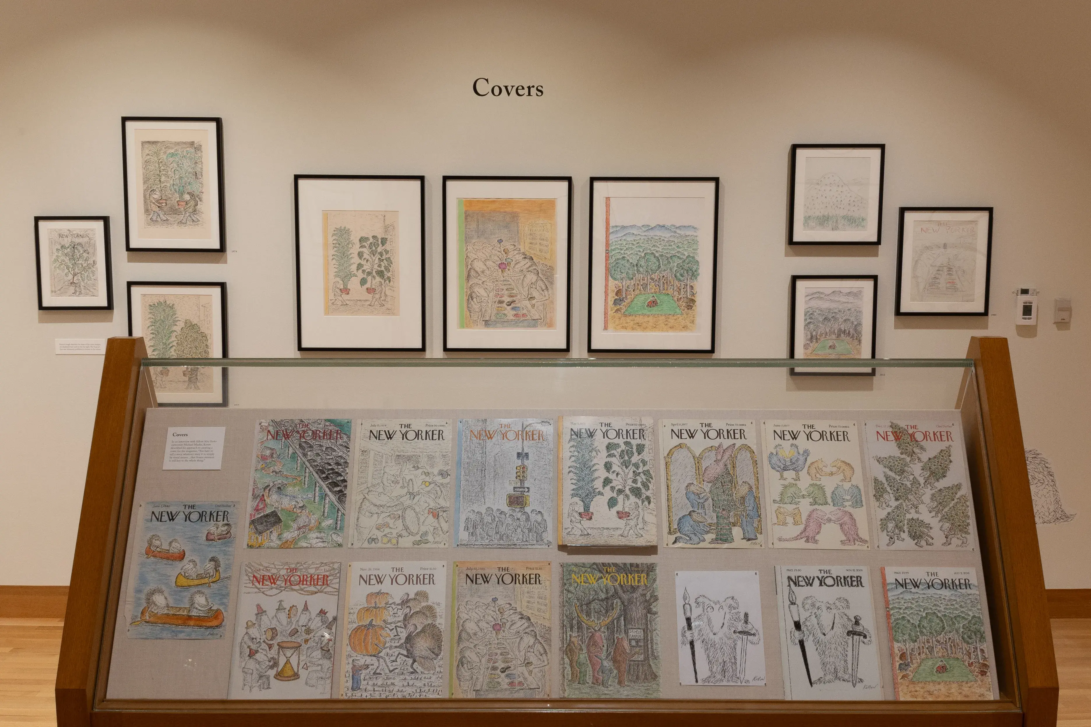 Installation image of a gallery exhibition. There are framed drawings on the wall and New Yorker magazine covers in a glass case in front.