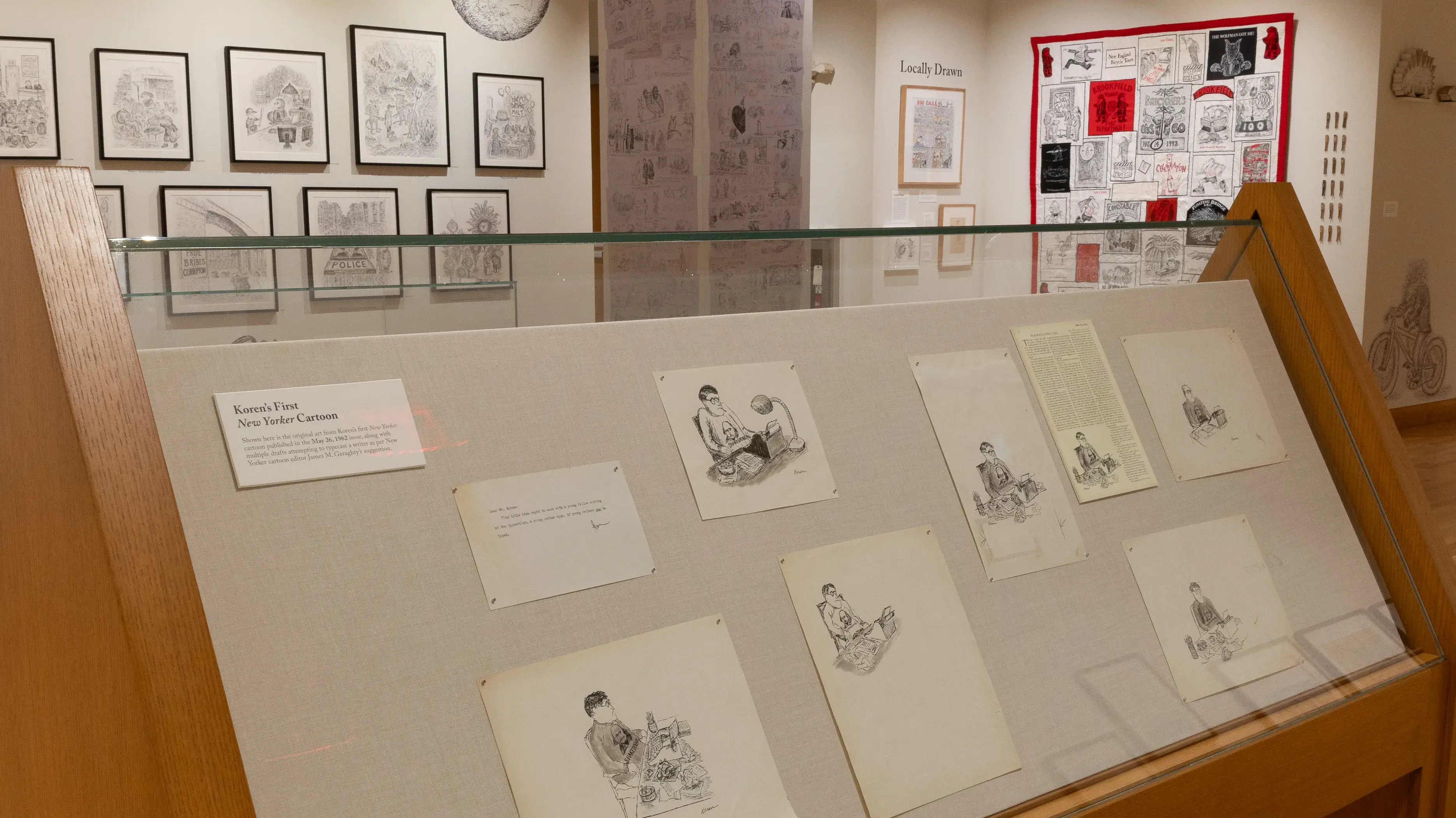 Installation image of a gallery exhibition. There are framed drawings on the wall and more drawings in a glass case in front. There are drawings on a column in the middle of the space and other objects including a quilt on the wall in the back.