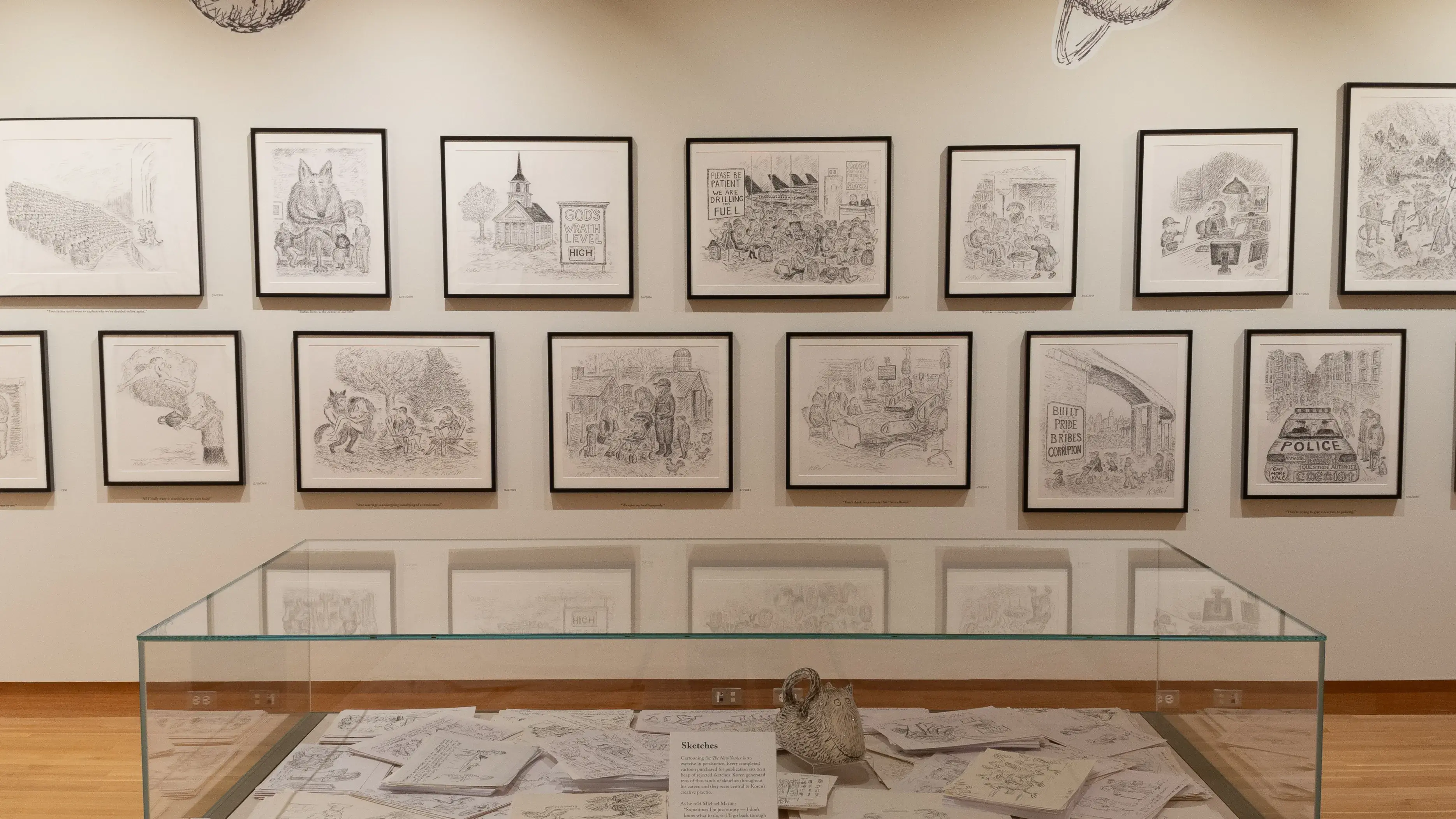 Installation image of a gallery exhibition. There are framed drawings on the wall and more drawings in a glass case in front. 