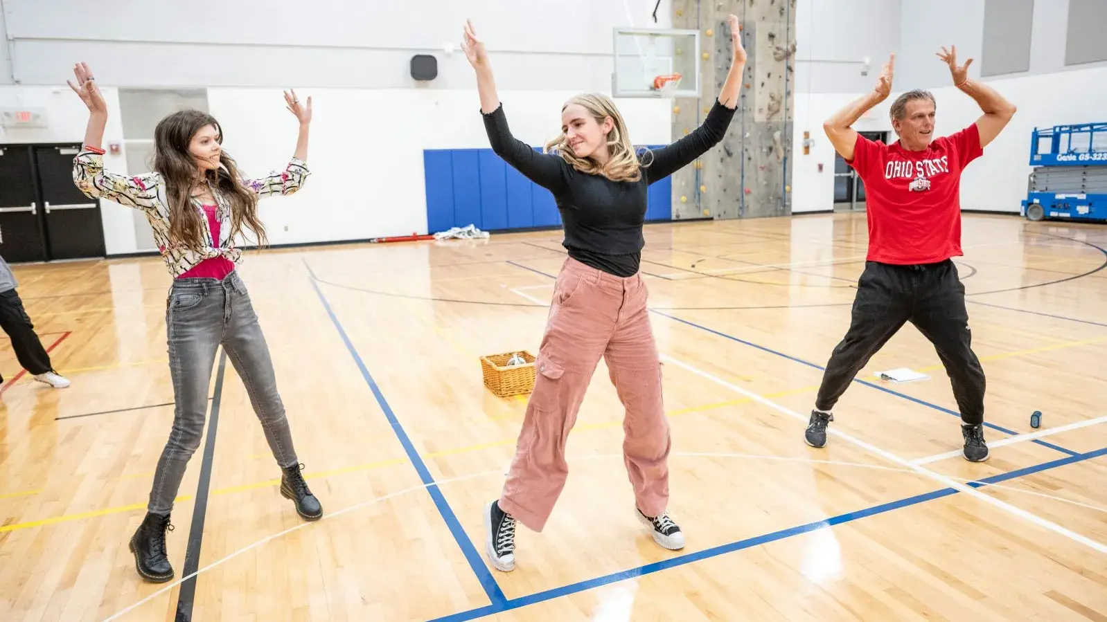 Two young people and one adult are standing on the wooden floor of a gym. All three look like they're jumping.