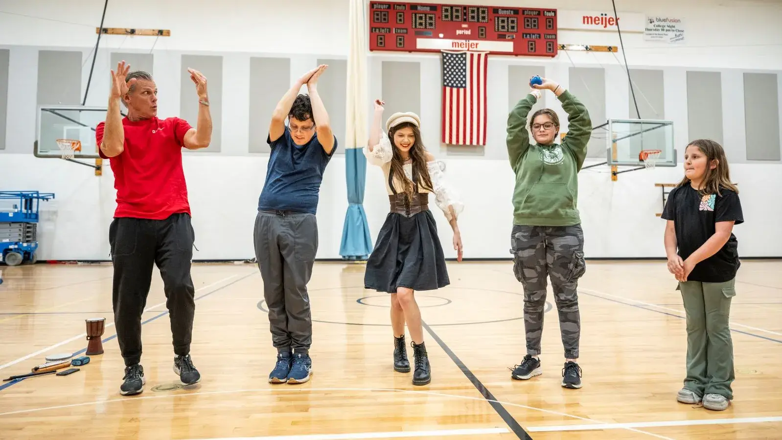 An adult and several young people stand on the floor of a gym and are moving their bodies in various ways