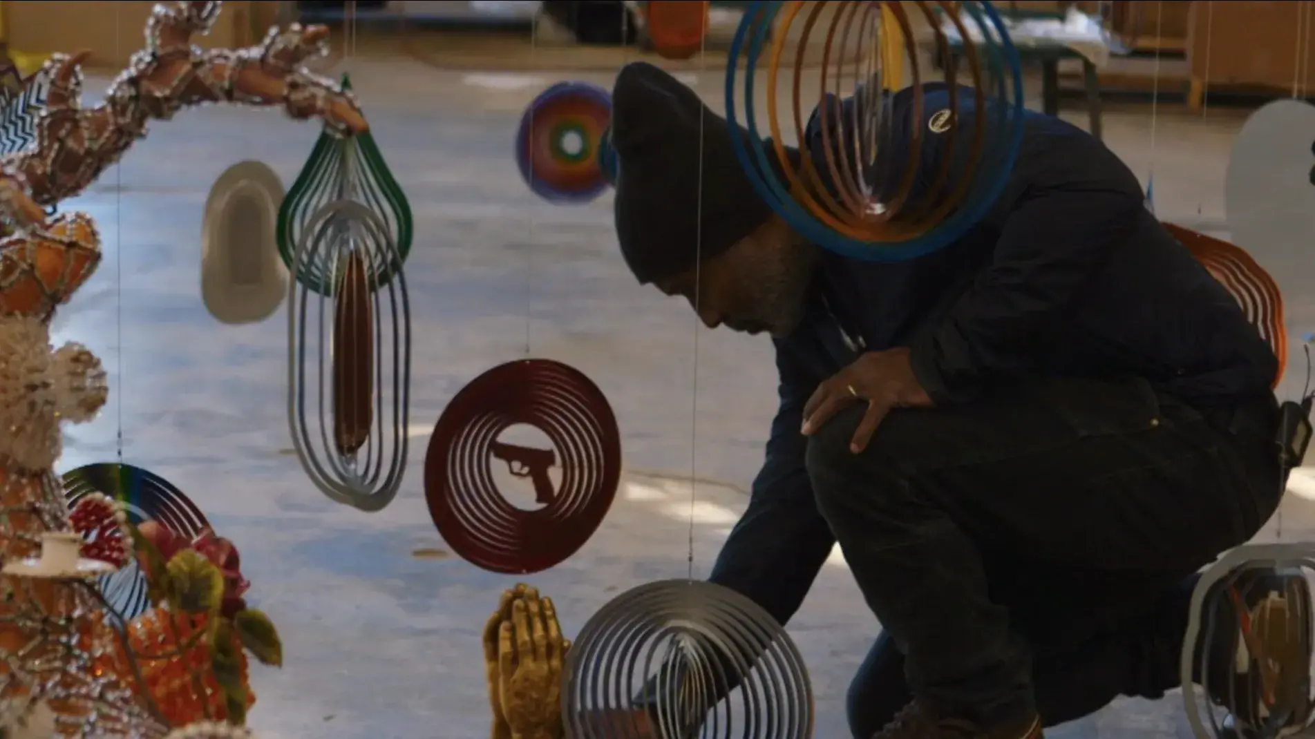 A man kneels on the floor surrounded by colorful hanging sculptures.