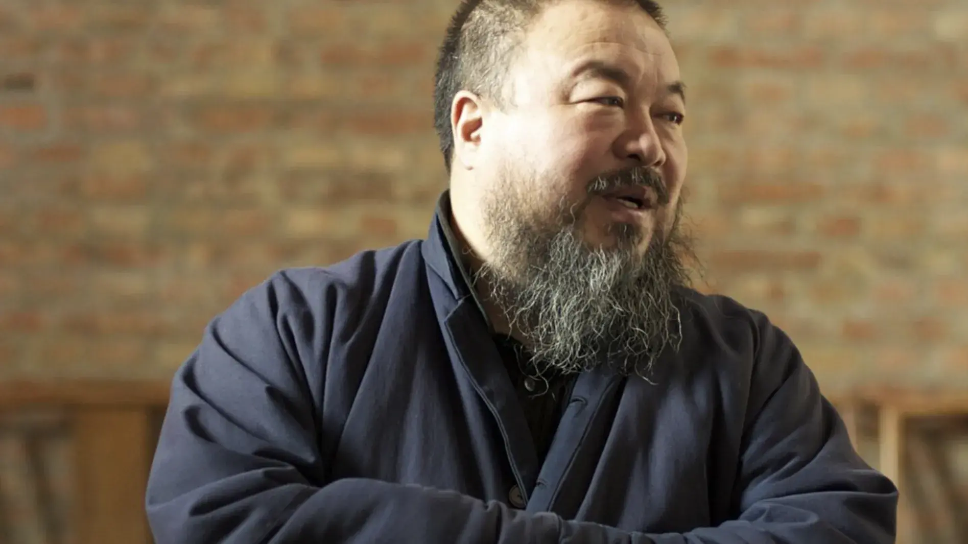 Photo of Ai Weiwei, a man with a beard wearing a blue jacket with his arms folded standing in front of a brick wall.