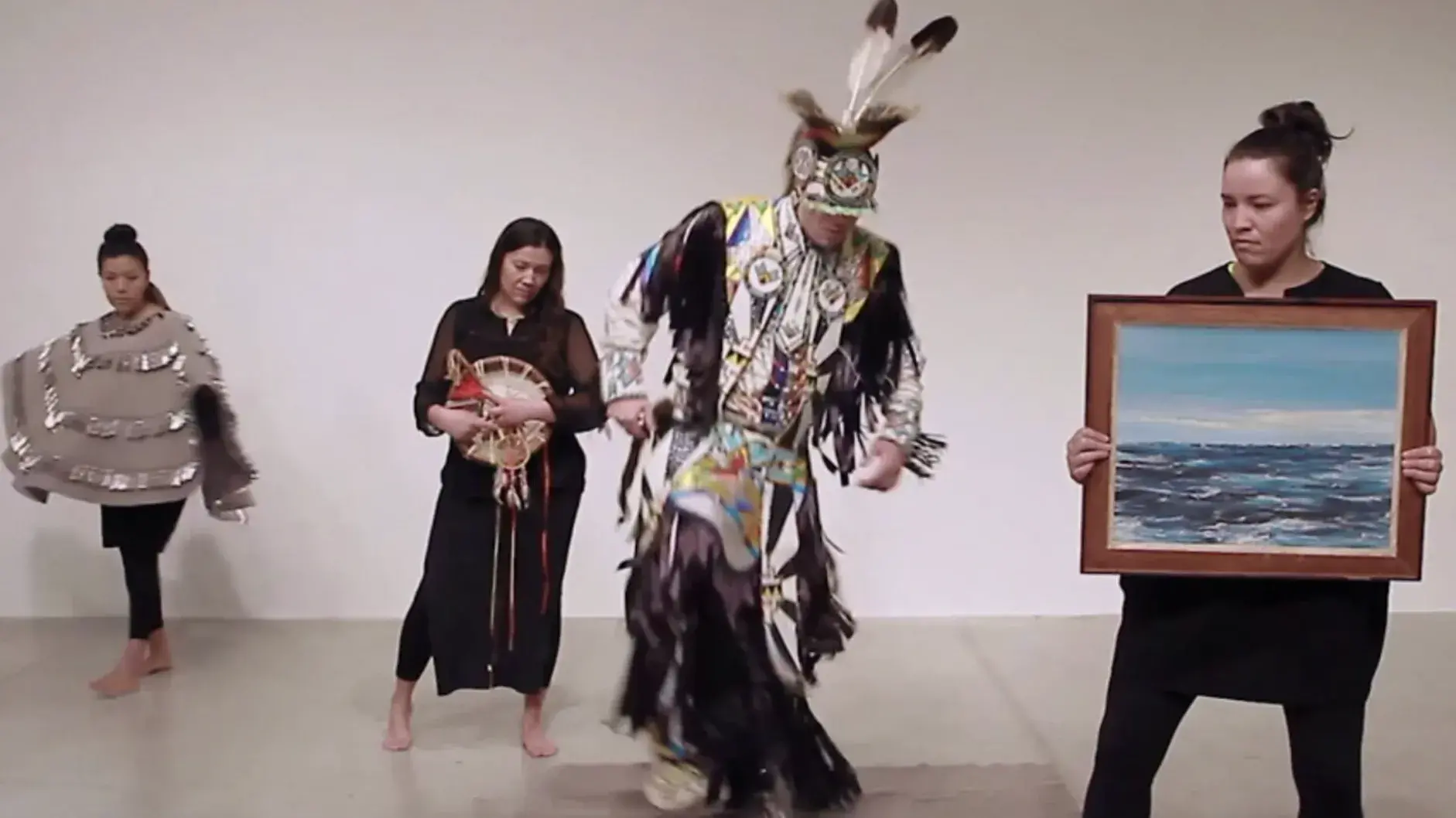 Four performers wear Indigenous ceremonial or black clothing. One wears a gray felt shawl with silver jingles. One holds a landscape painting.