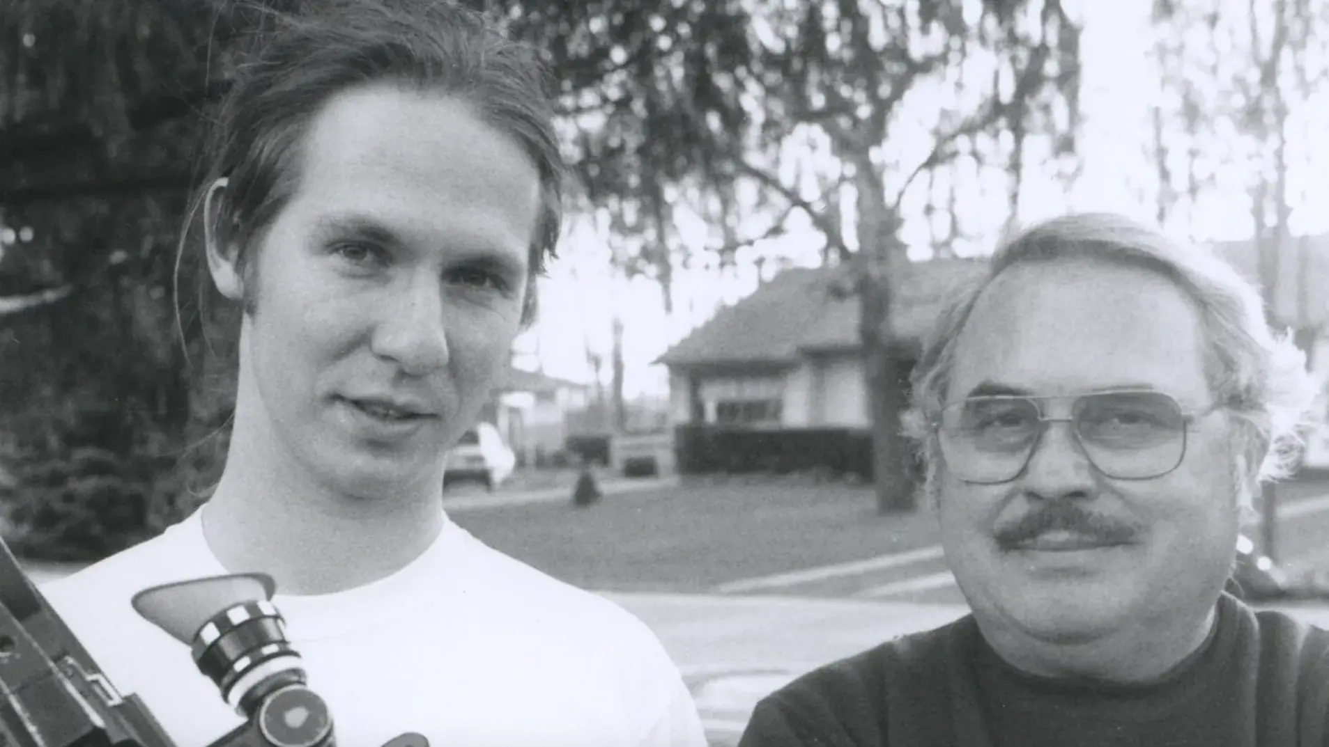  Black and white image of a young man holding a film camera standing next to an older man whose arms are crossed.    