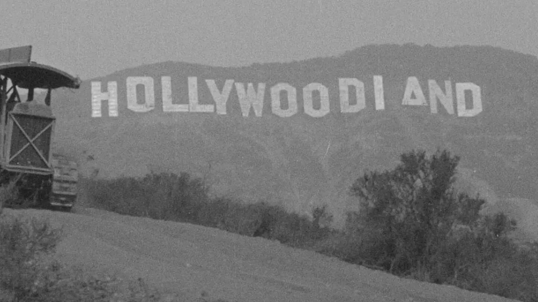 Black and white image of the original Hollywoodland sign on a hillside, partially obscured by a hill and bushes, with a vehicle nearby.    