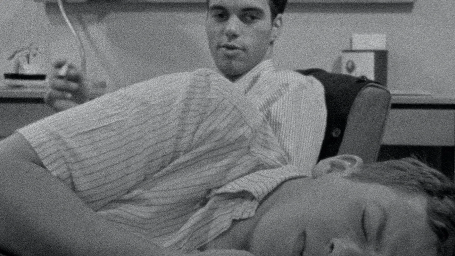 A young man is laying down and another young man is sitting behind him holding a cigarette. A desk, lamp, and bulletin board are in the background. 