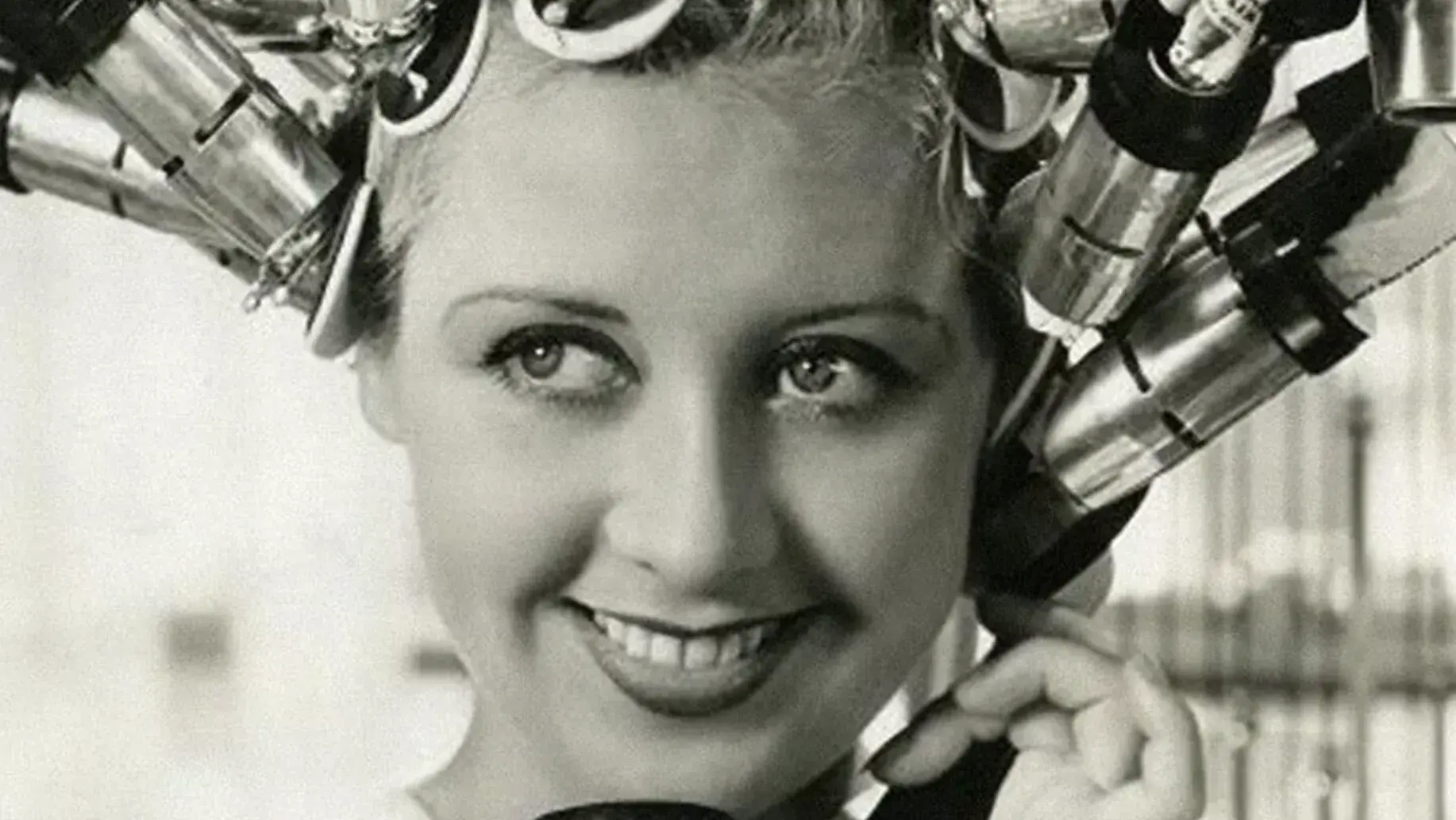 Black and white image of a smiling woman with her hair up in a large hair curling machine. 