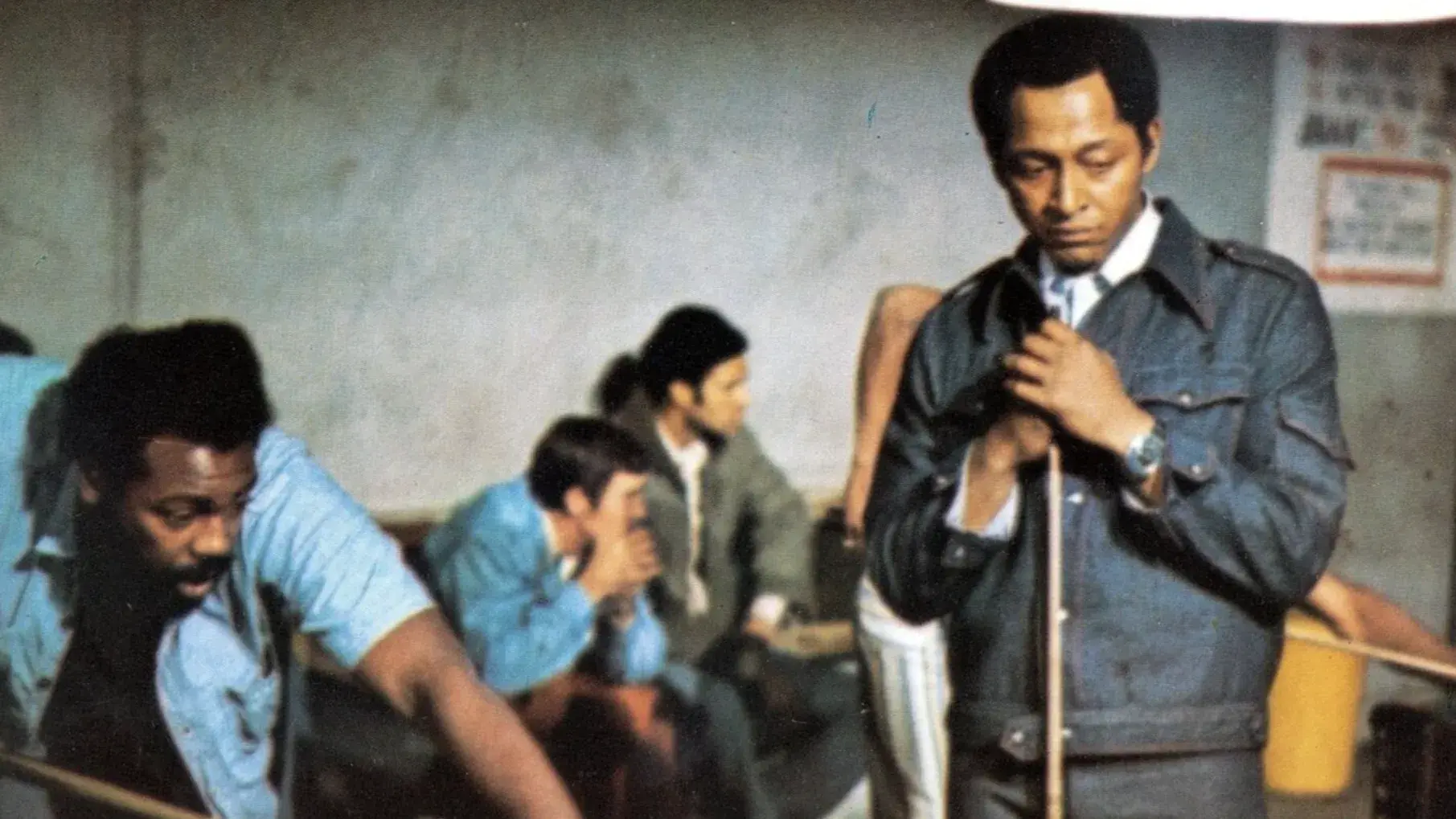 Two Black men playing pool in a pool hall. One man is bent over shooting, the other is standing nearby watching. 