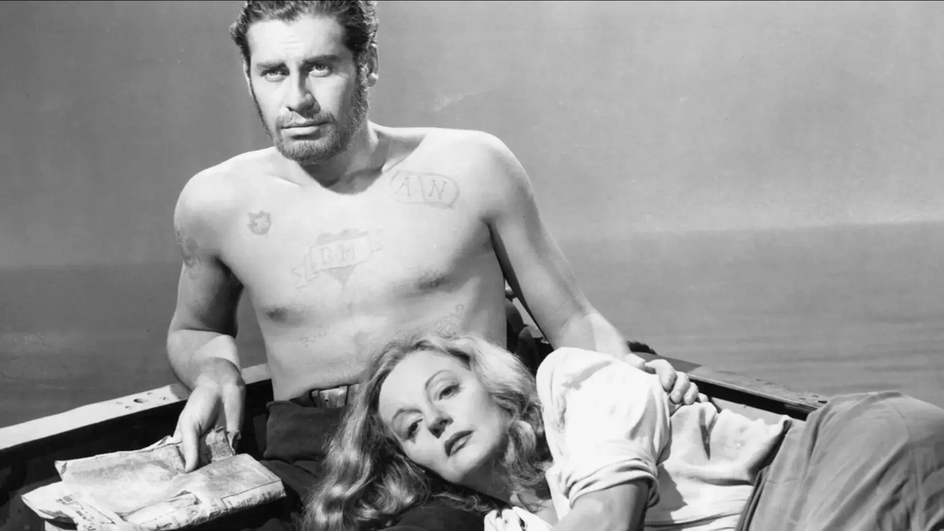 A man sitting on a small boat with a woman resting her head in his lap. They are surrounded by water. 