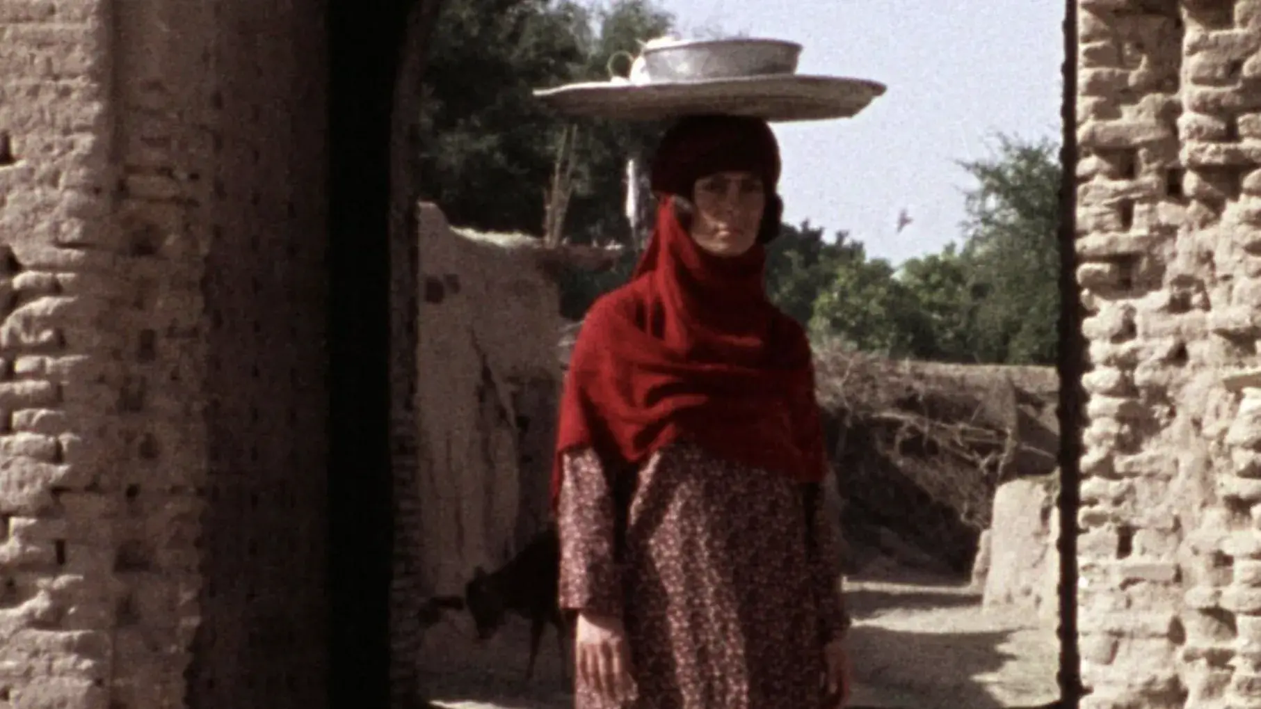 A woman is standing under the arch of a very old looking building. She is balancing a tray and bowl on her head. 