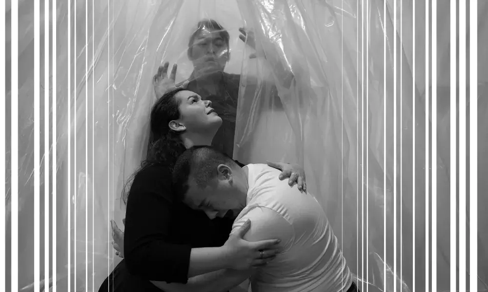 Image of a man and woman embracing and another man stands behind a clear sheet of plastic behind them.