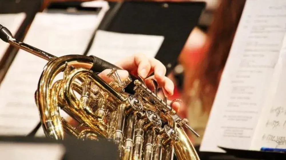 Image of a brass instrument being played with sheets of music on music stands in the background.