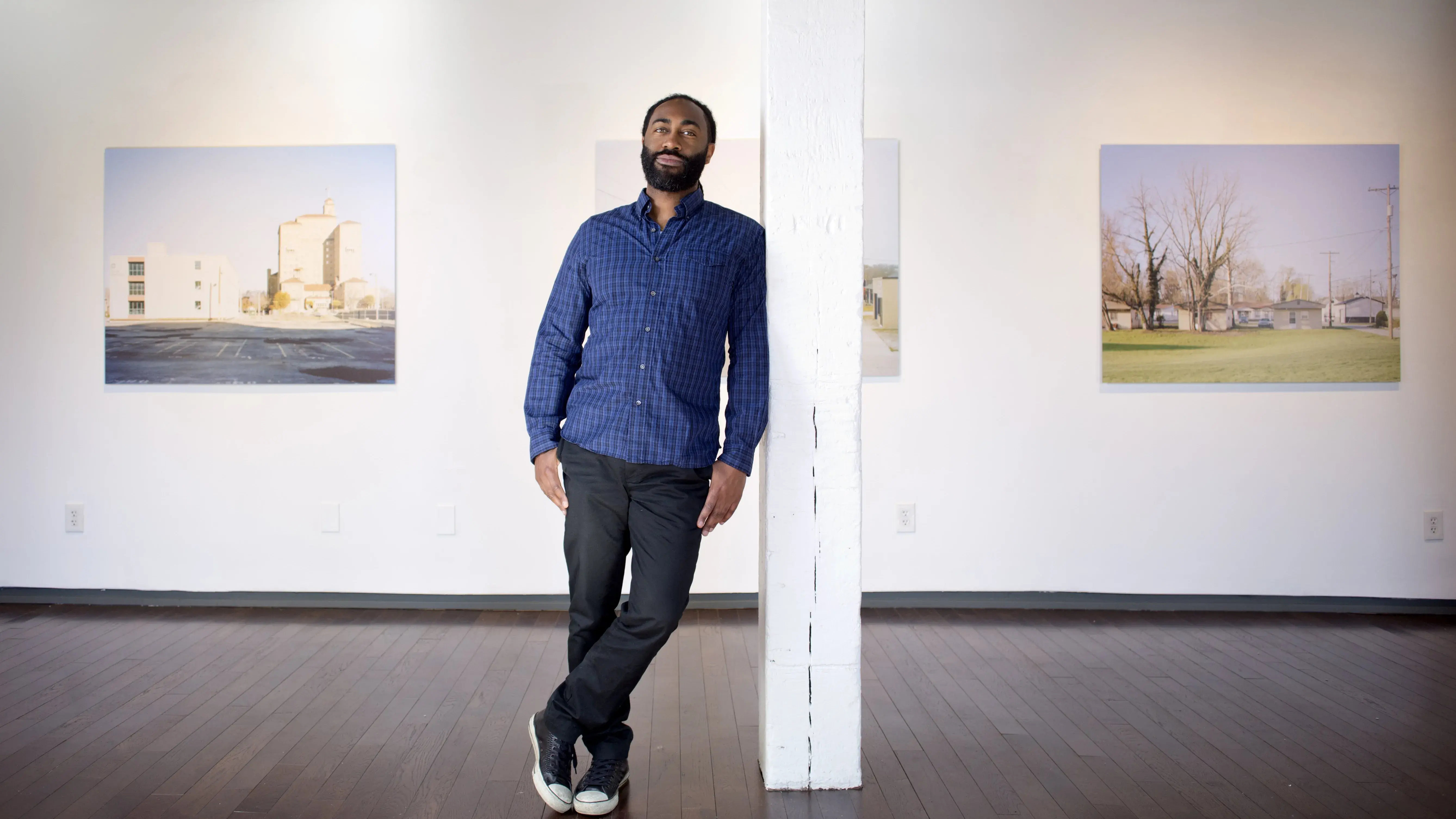 A black man with a trimmed beard leans against a white pole. Several large photographs of buildings are hung on the wall behind him.