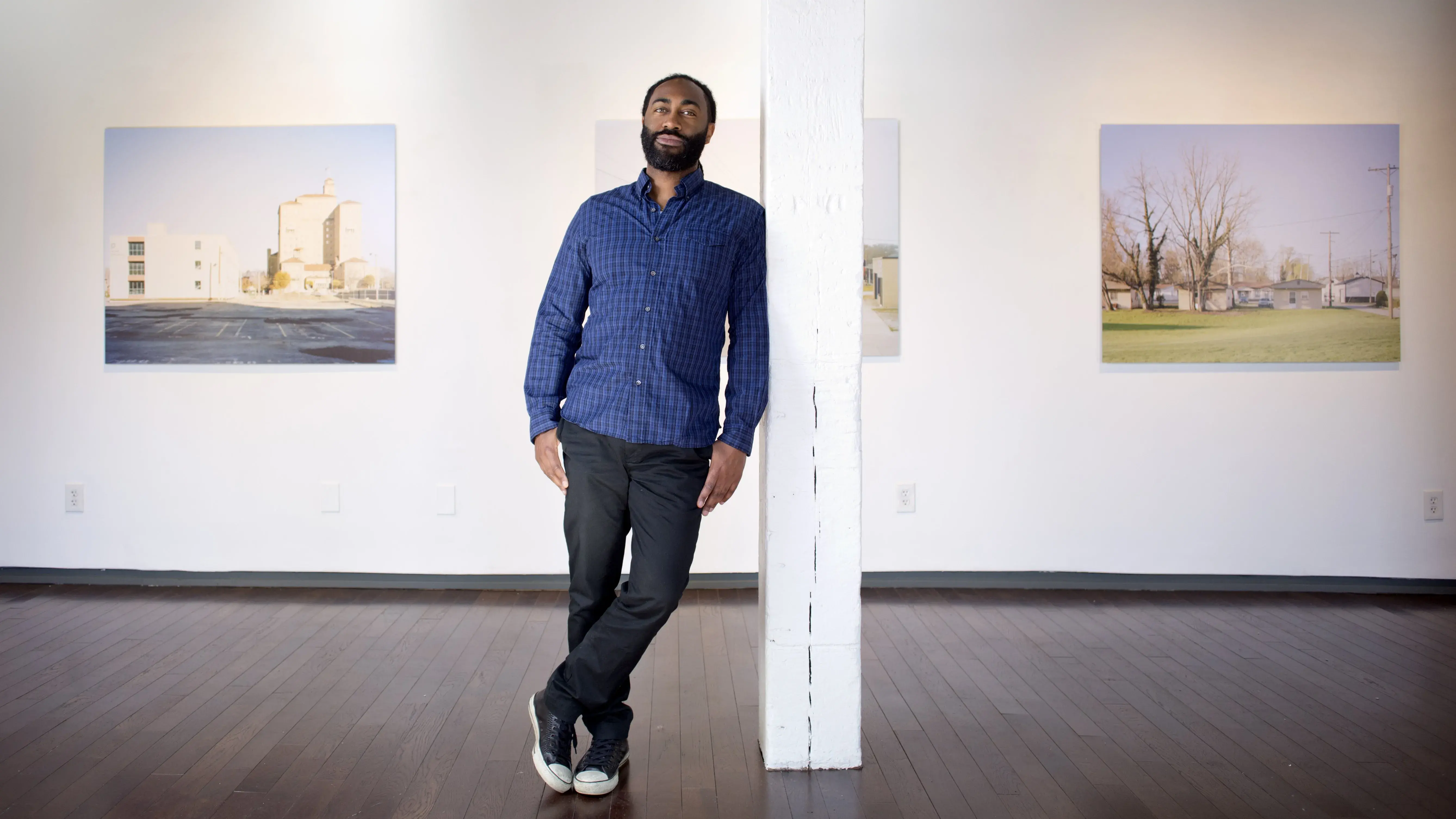 A Black man with a trimmed beard leans against a white pole. Several large photographs of buildings are hung on the wall behind him.
