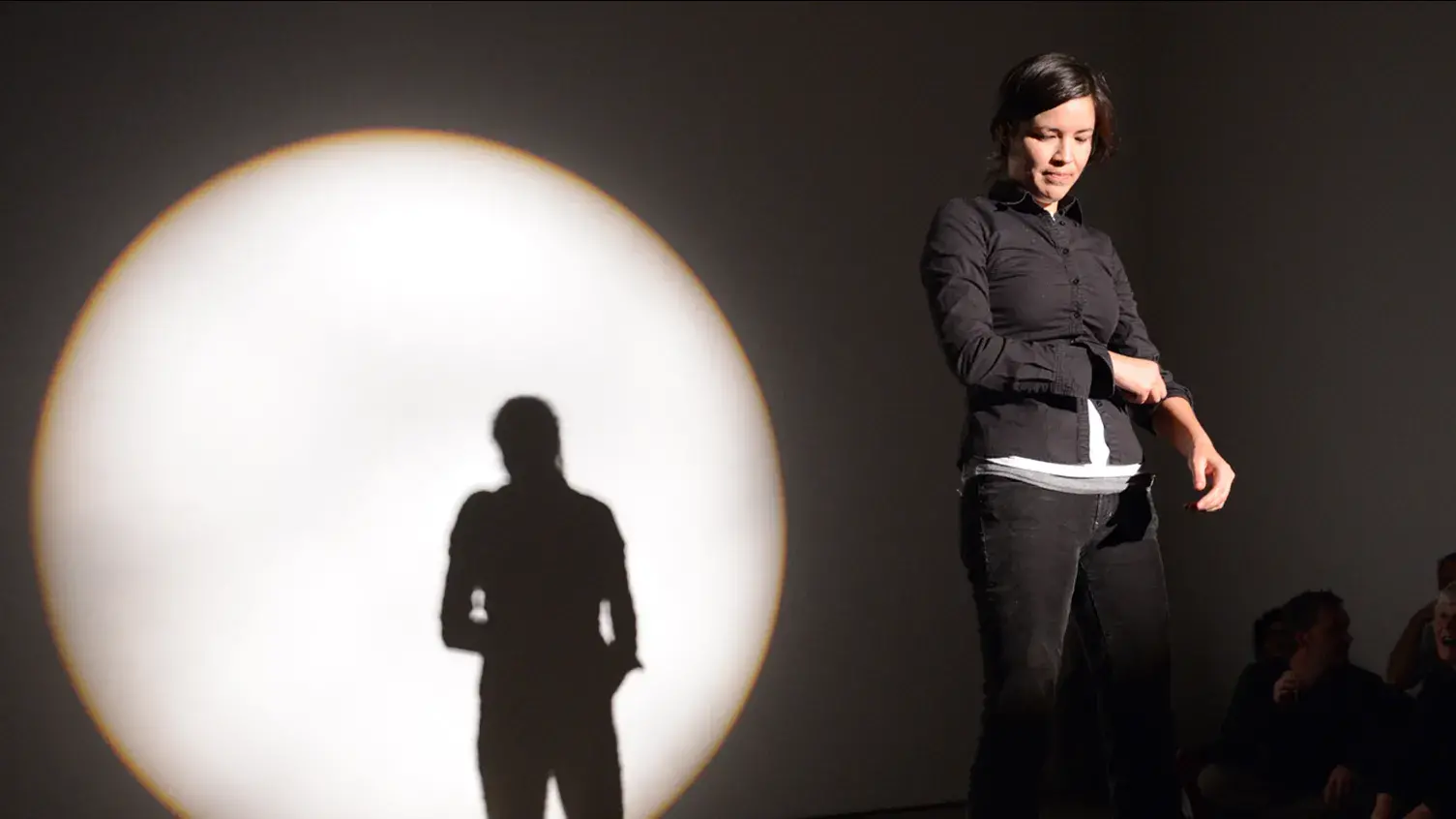 Maria Hupfield stands in a spotlight onstage wearing a black button-down shirt and jeans. Her silhouette is visible in a large circle of light behind her.