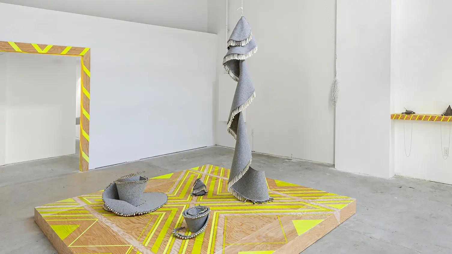A wooden plinth with a yellow geometric pattern supports three gray felt and jingle sculptures; a fourth hangs and spirals down onto the platform.