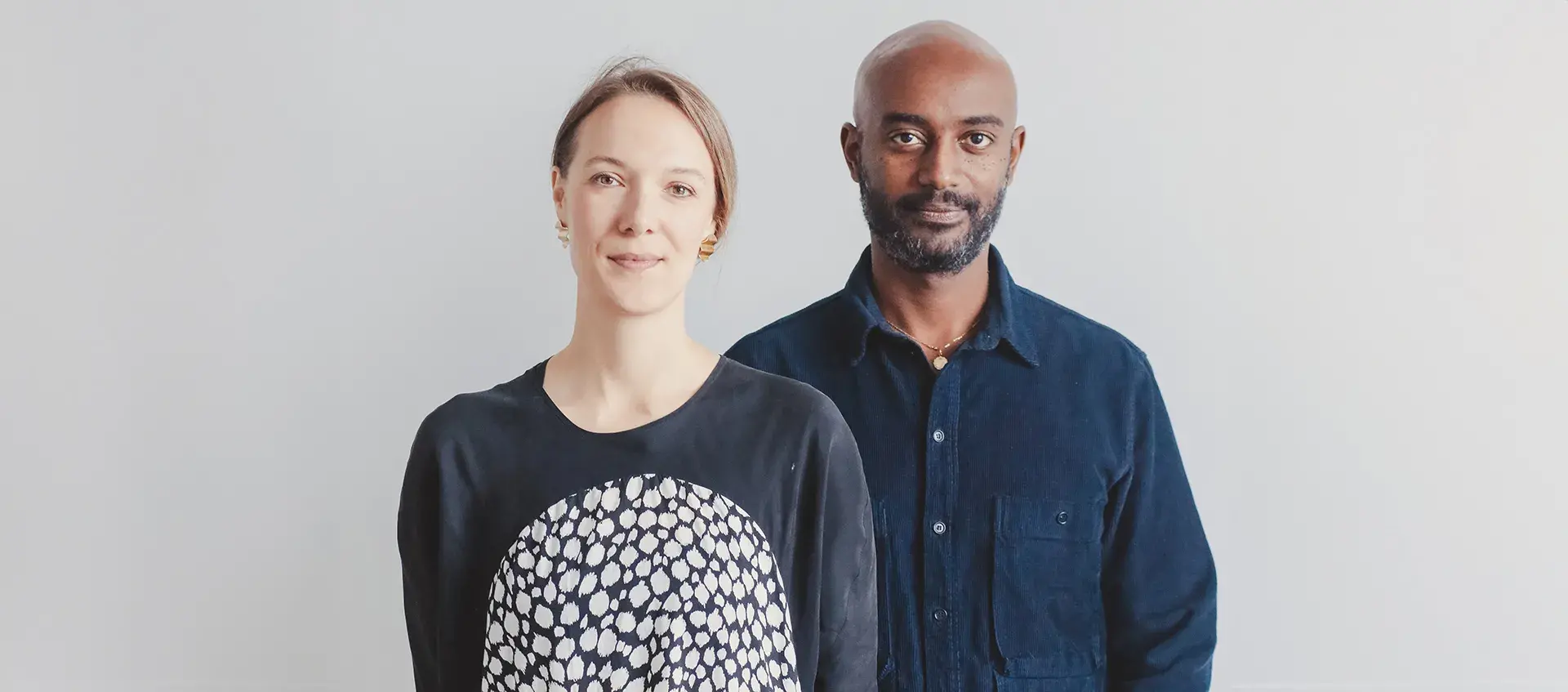 Jen Wood, who has pale skin and light-brown hair stands to the left of Emanuel Admassu who has medium brown skin, is bald, and has a short beard.