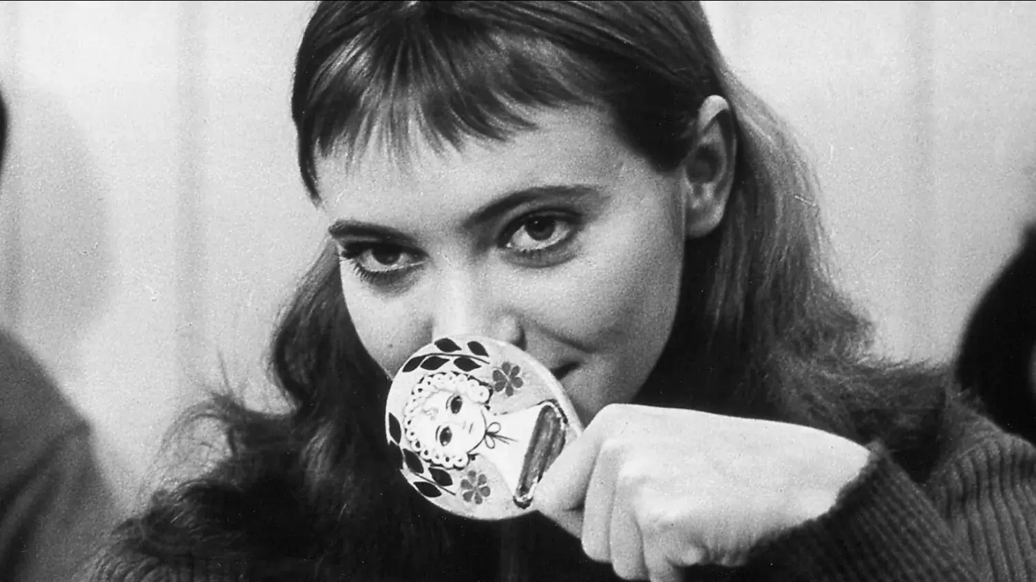 A young woman with bangs holds a hand mirror up to her nose with a painting of a woman wearing a bonnet on the back.