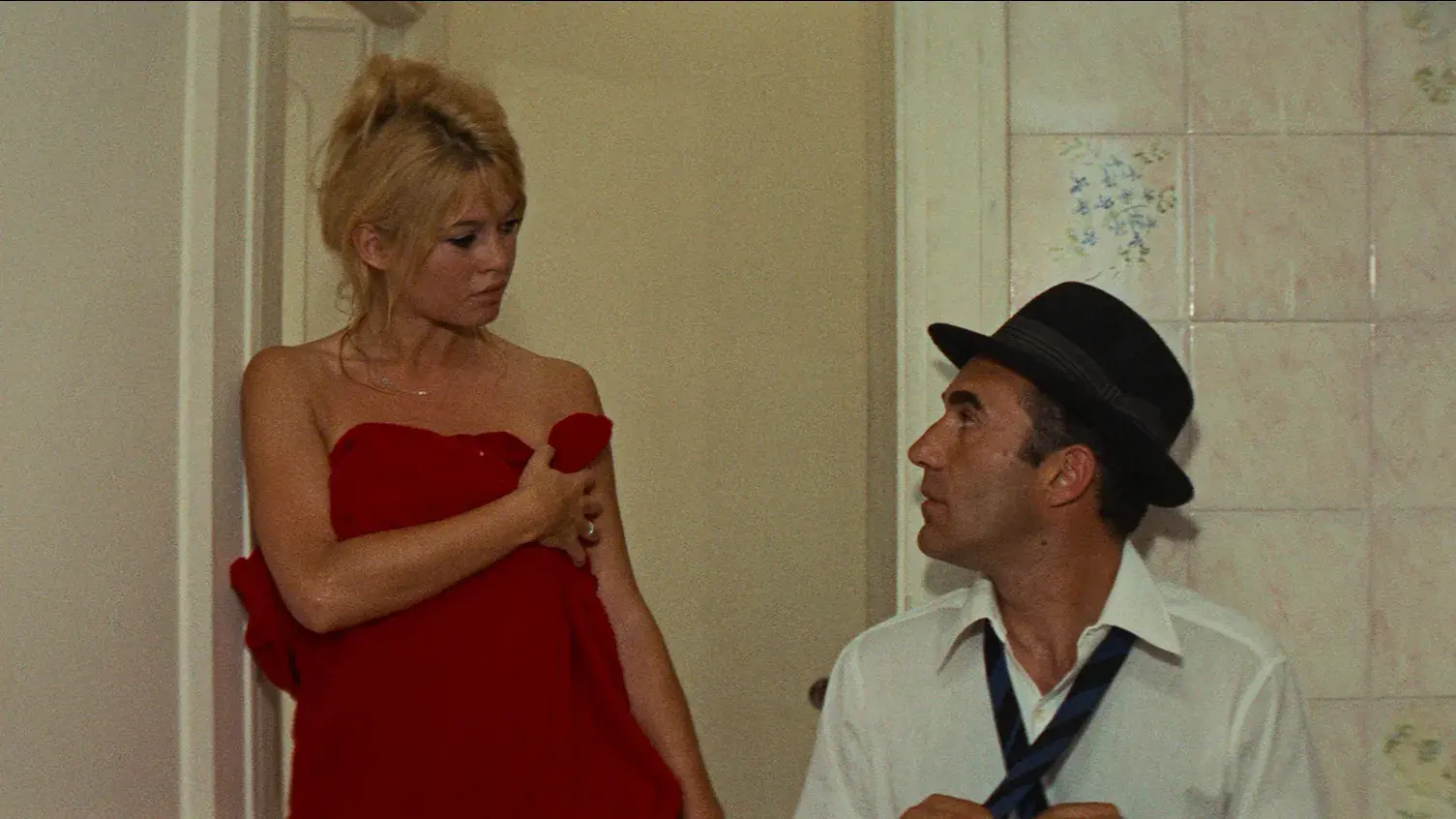 A woman wrapped in red towel is leaning in a doorway next to a man seated next to her wearing a fedora and tying a tie.