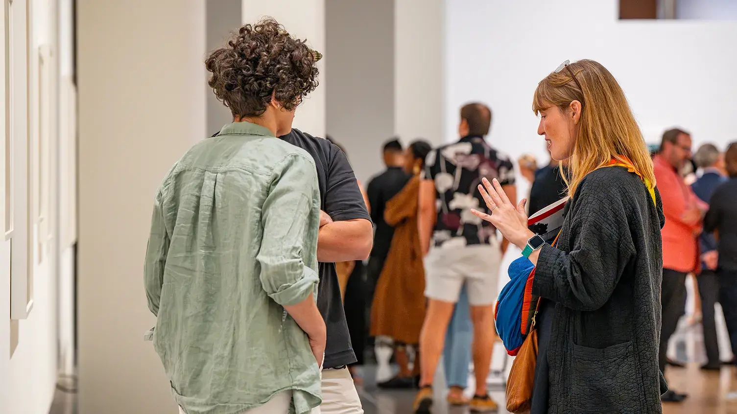 People stand in groups in a large gallery. In the foreground, three people have a conversation next to framed works hanging on the wall.