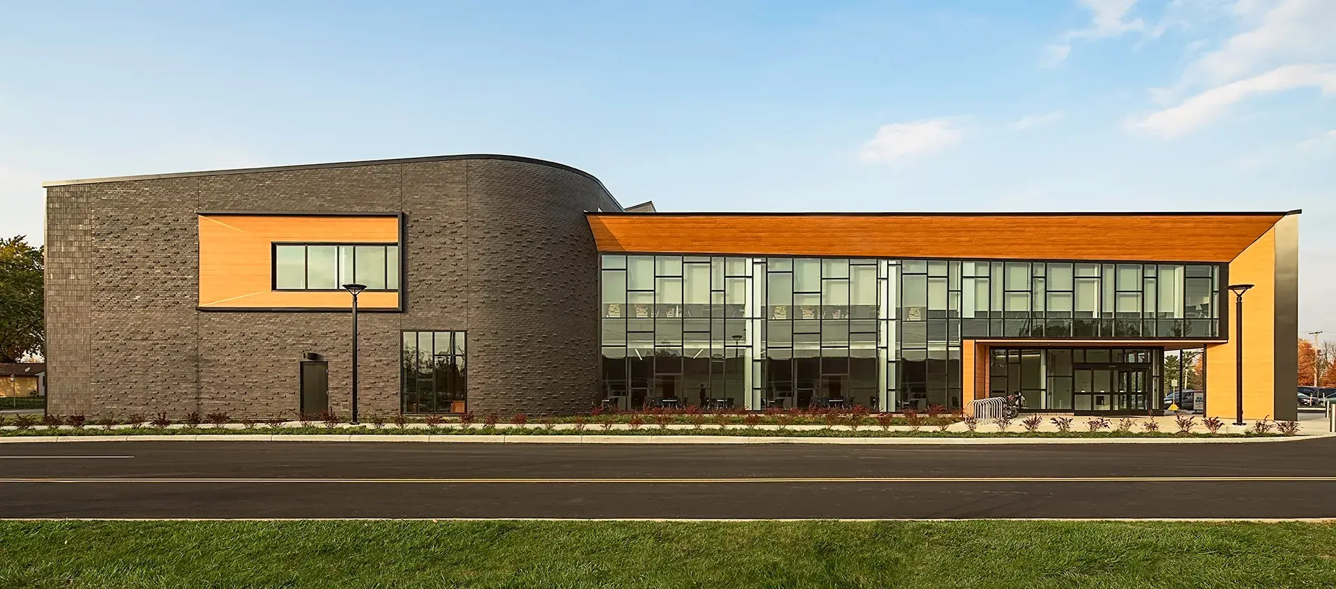 A street view of a building. A taller brown, brick, cube with curved corners intersects with a long wood-framed rectangle featuring a wall of windows.