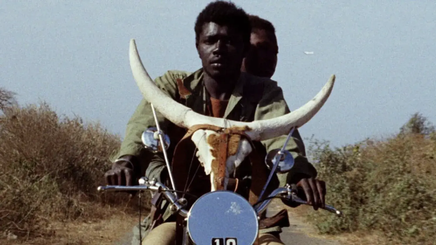 A black couple ride a motorcycle with a horned skull on the handlebars through a desert landscape. 