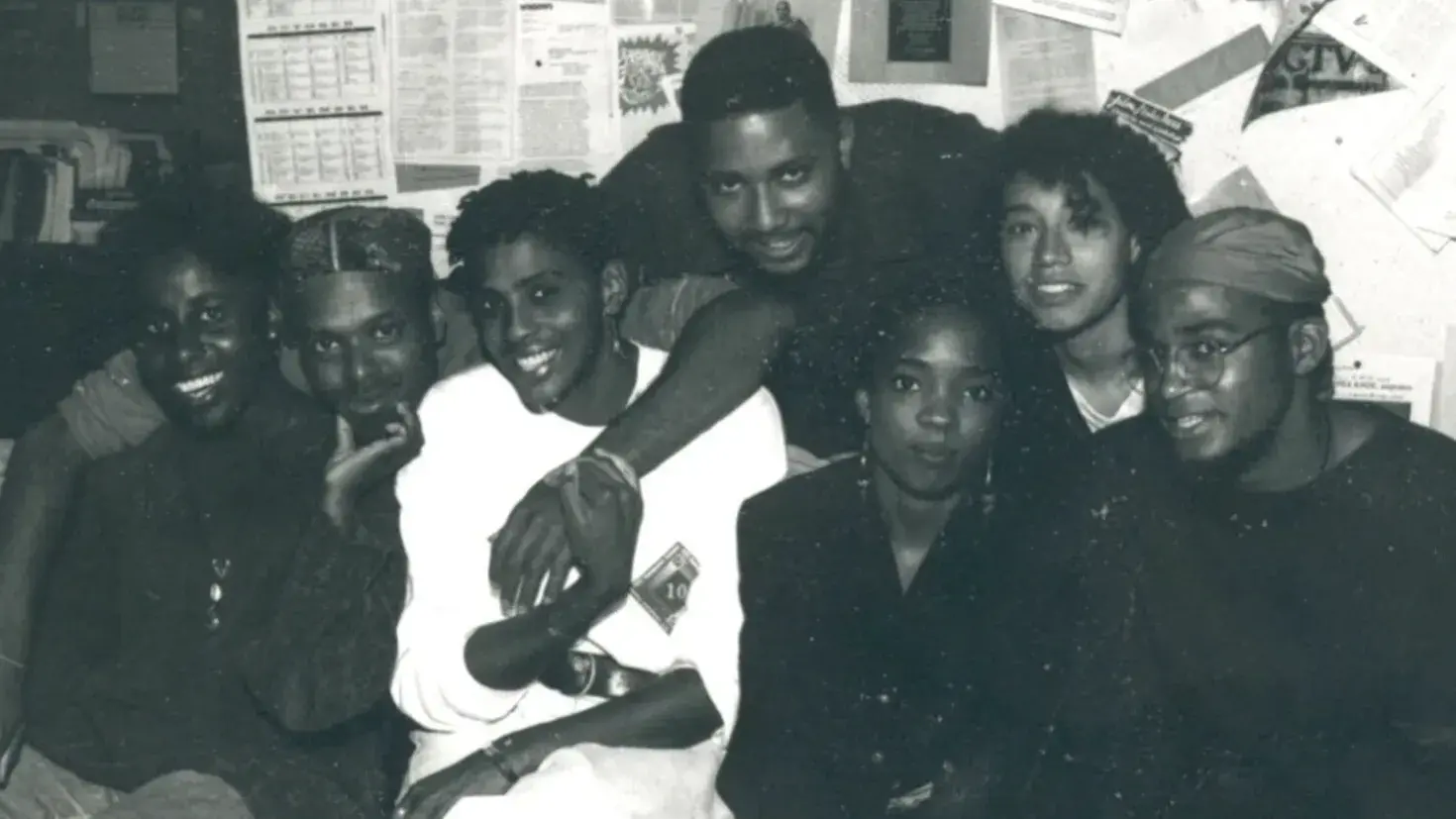A group photo of seven young, Black individuals, they are smiling with their arms around each other. 