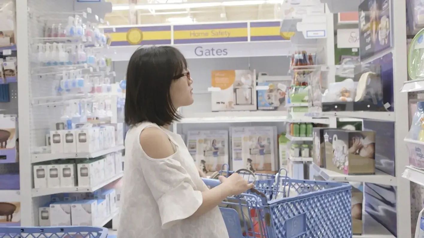A pregnant Asian woman pushes a shopping cart through the baby section of a store. 