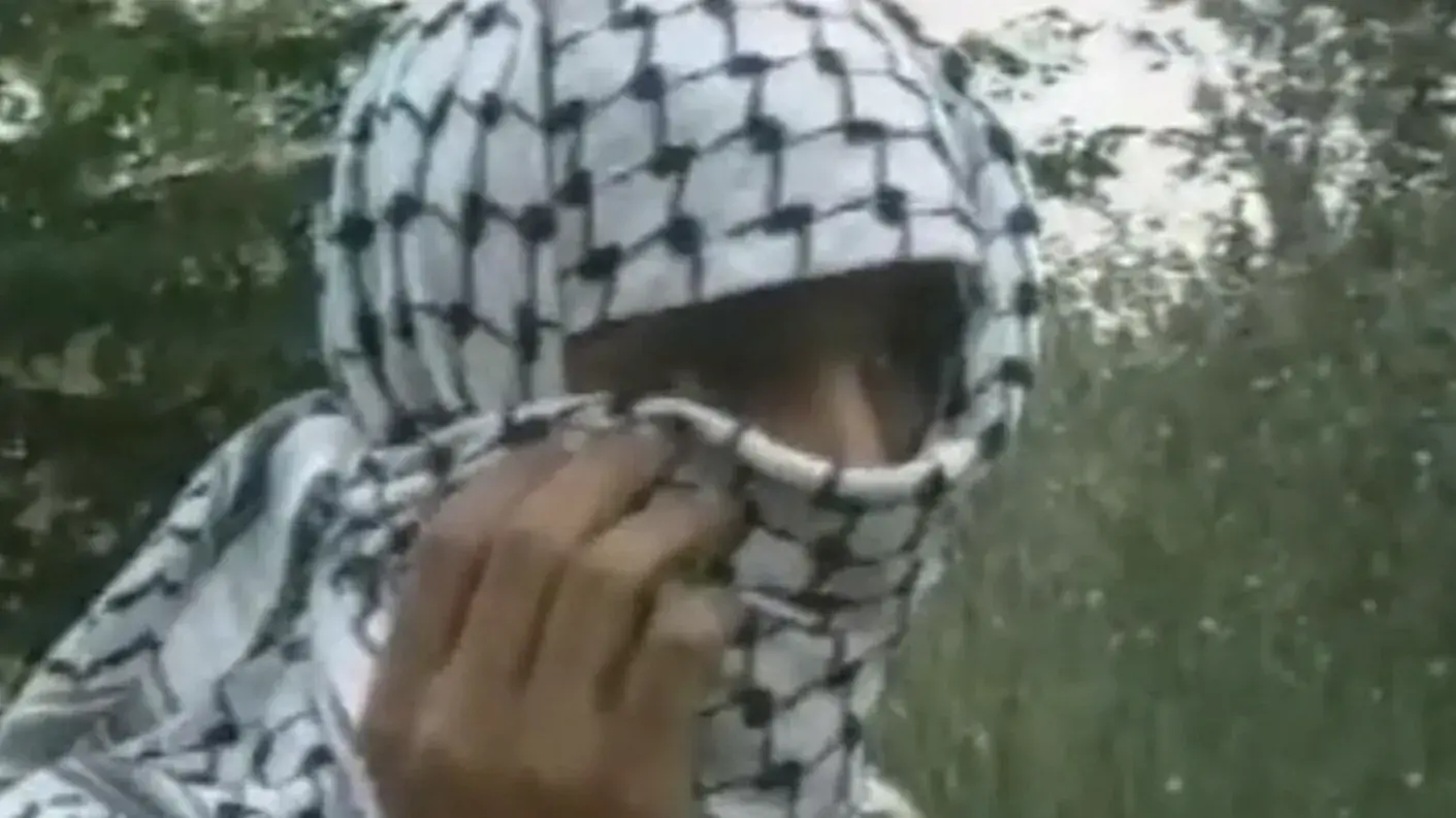 A man wearing a kaffiyeh over his head and face standing in front of trees. 
