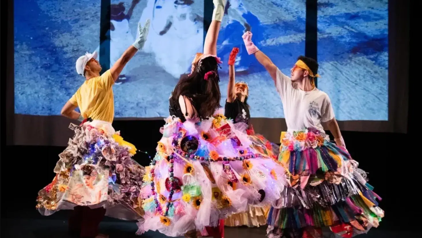 Four people wearing big multicolored skirts reach into the air in front of a blue projected background