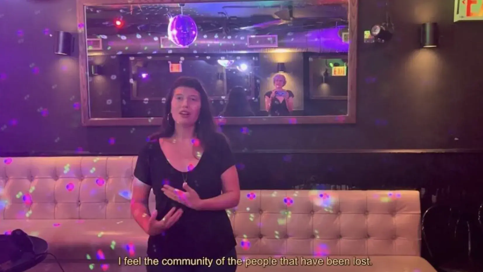 A person in a black outfit stands in front of a shiny leather couch, above which is a mirror reflecting a person holding a camera.