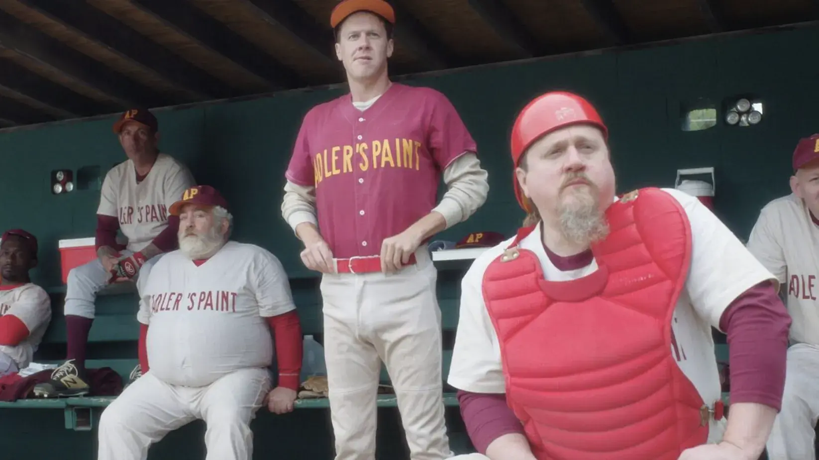 A baseball team sits in a dugout, the men are older and one is wearing a catcher's uniform. 