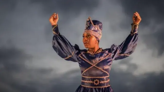 Black woman dressed up in a purple piece with a crown on her head with arms up. 