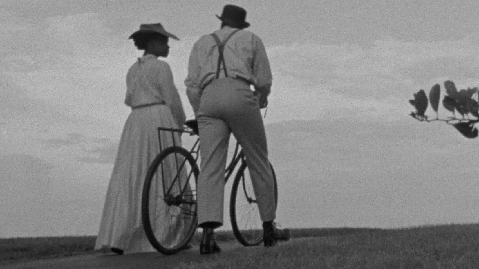 A black and white image of a man and a woman, dressed in vintage clothes, walking on each side of a bicycle along a country road. 