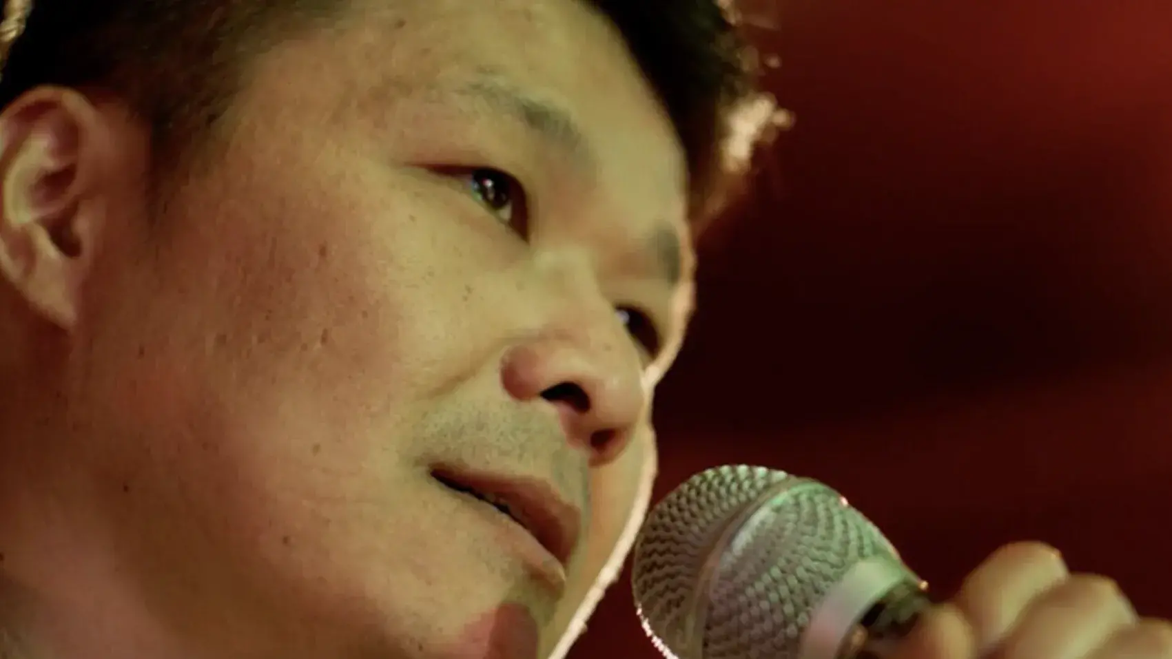 A man stands in front of a red background holding a microphone. 