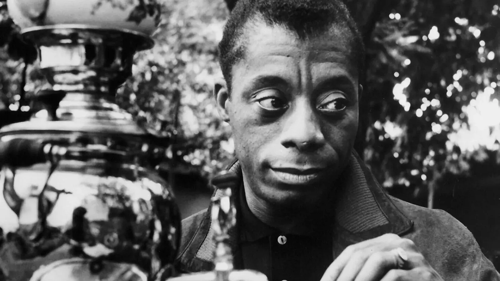 James Baldwin drinking tea, he is sitting outside next to an ornate large metal decanter. 