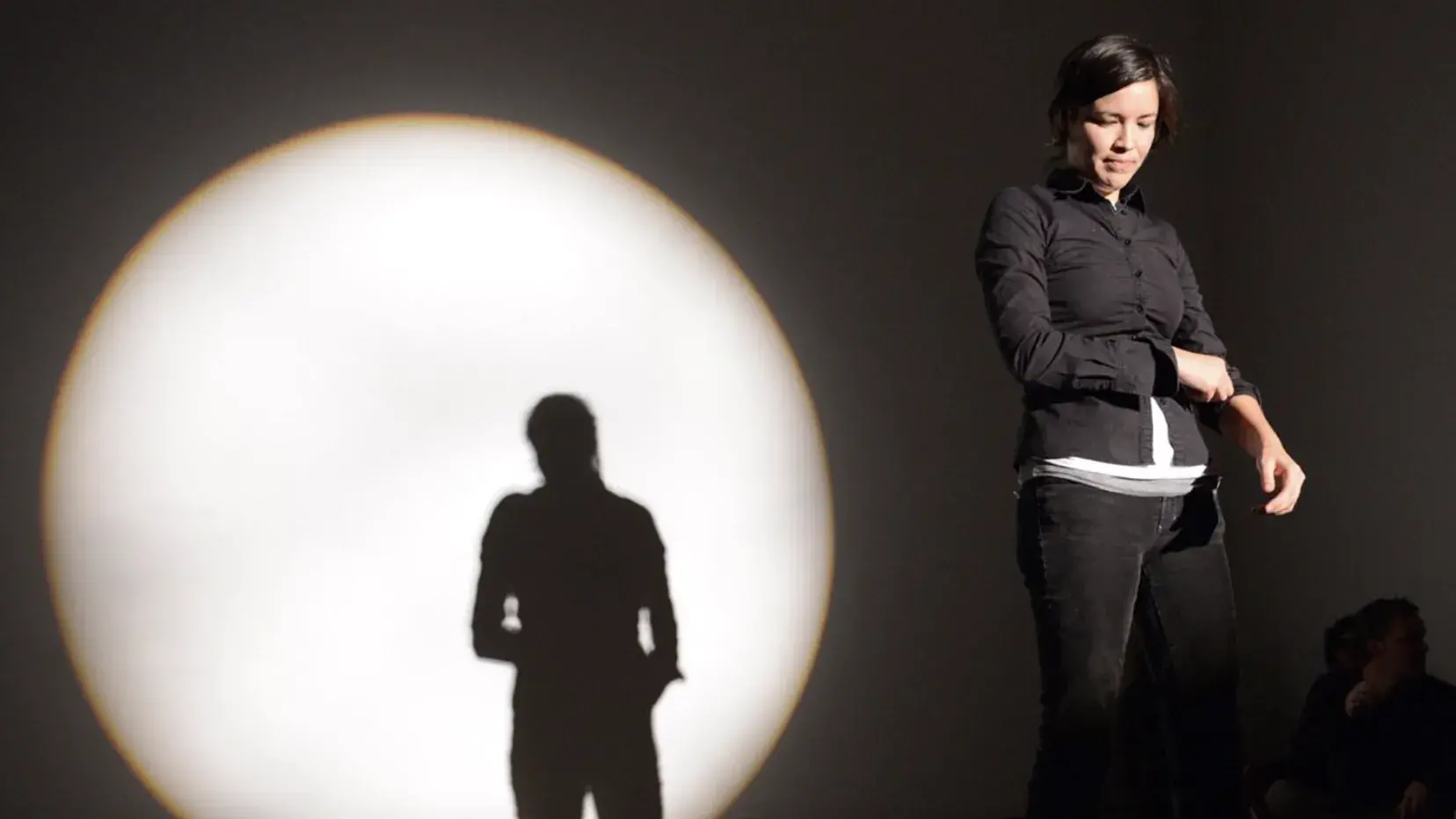 Maria Hupfield stands in a spotlight onstage wearing a black button-down shirt and jeans. Her silhouette is visible in a large circle of light behind her. 