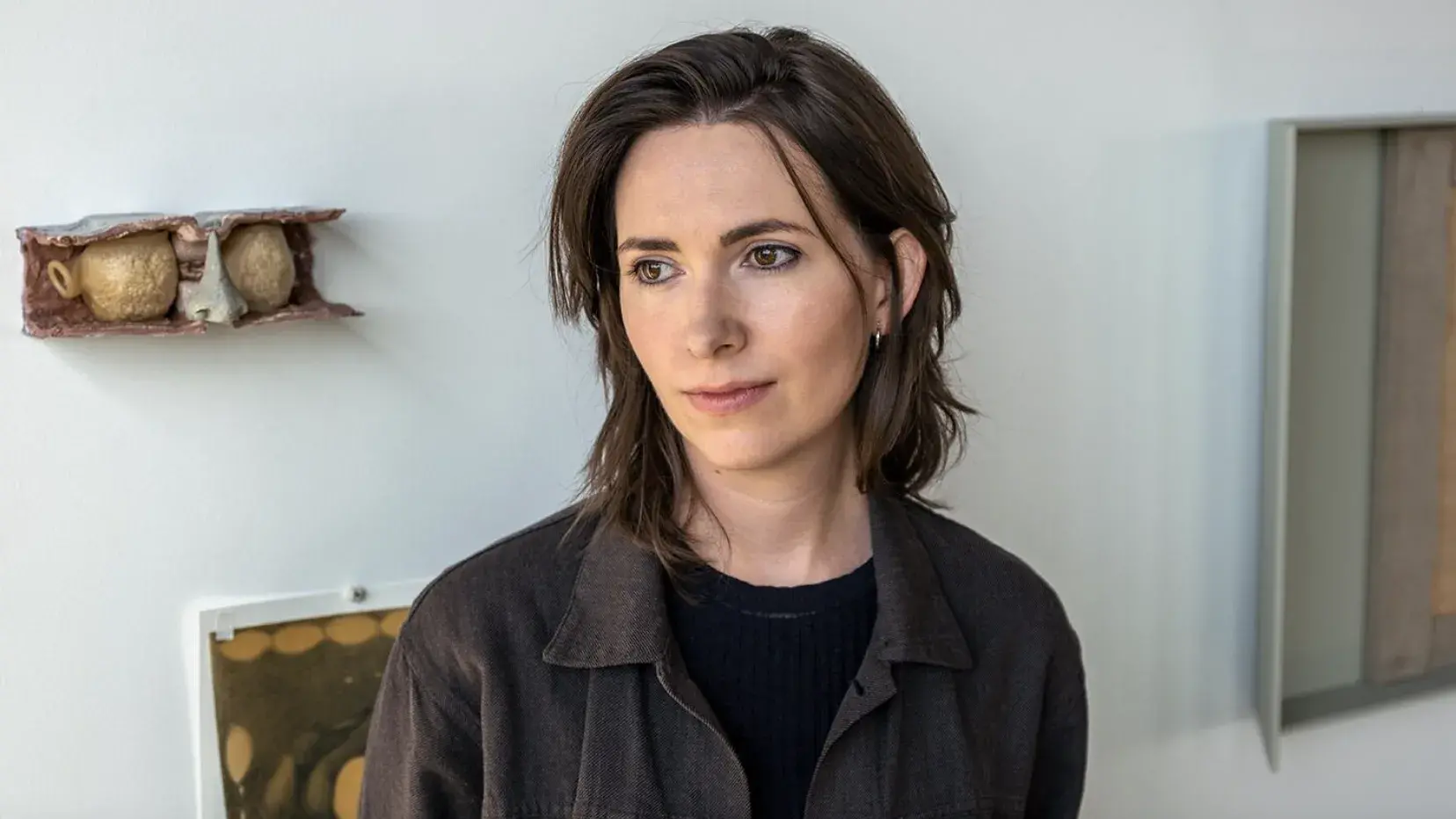 B. Ingrid Olson stands in front of a wall displaying her artwork. She has medium length brown hair and wears a gray jacket and black shirt. 