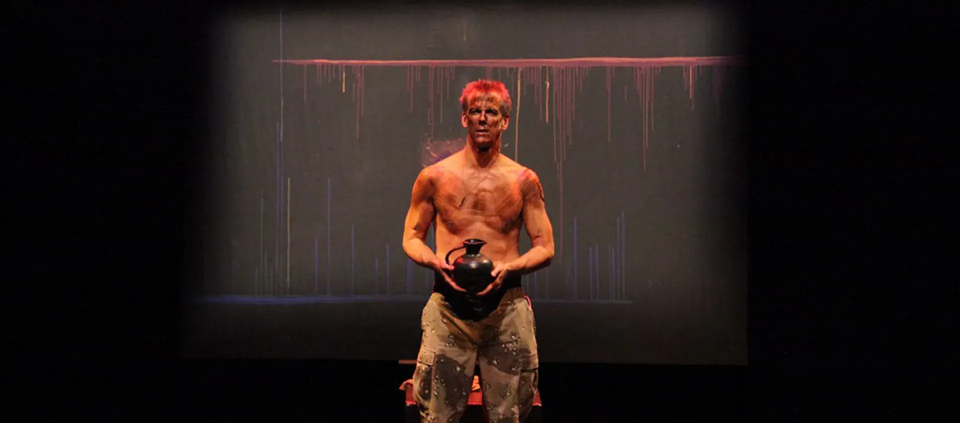 Kevin McClatchy stands holding a black pitcher in a dark space. He isn't wearing a shirt and has camoflage pants on. There are marks and colors on his face and torso and his hair is dyed bright red.