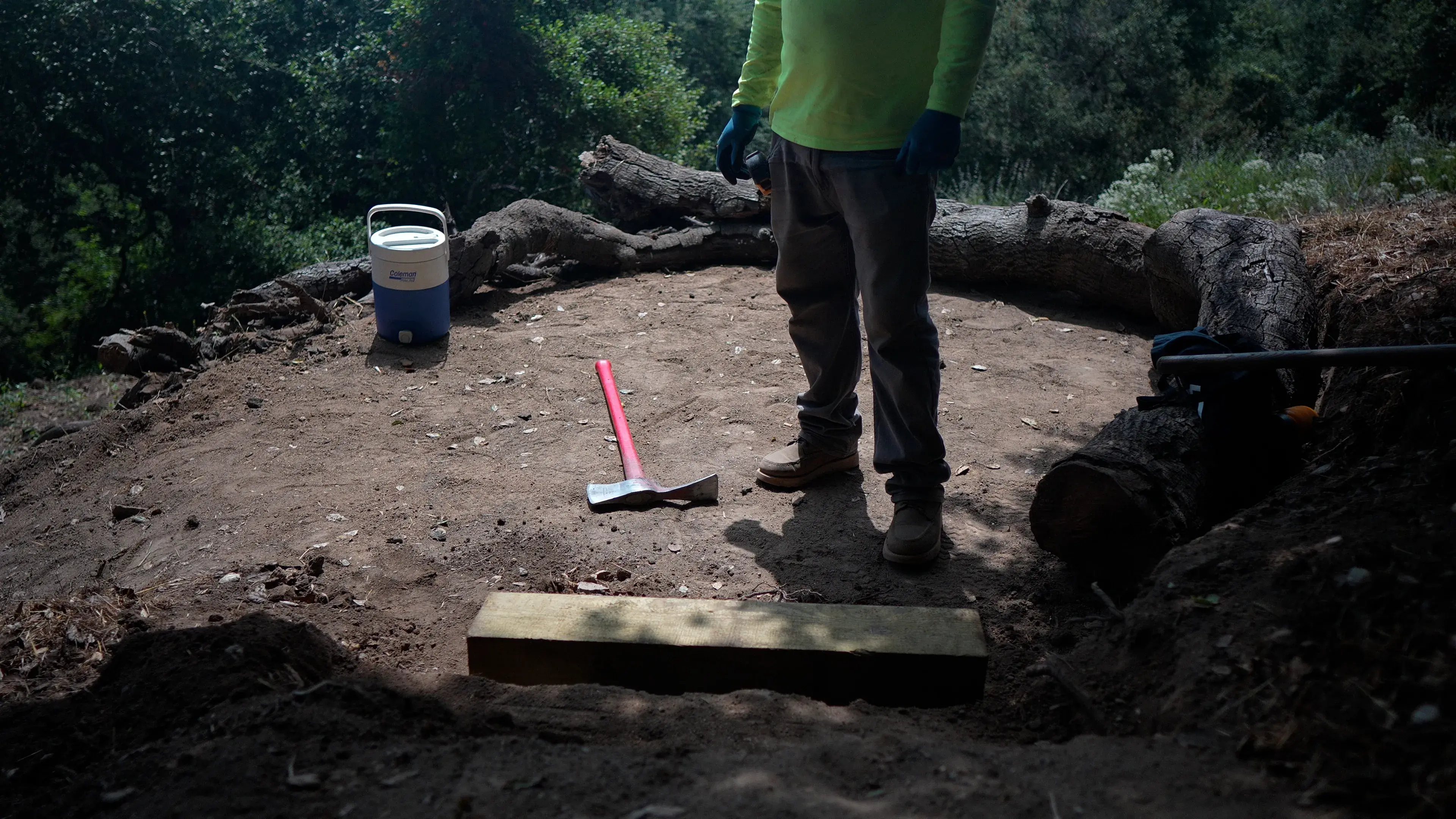 A person stands on dirt with an ax and a cooler on the ground next to him