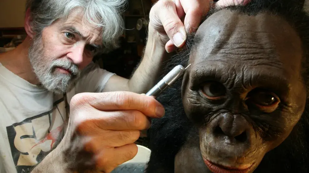 John Gurche, a man with gray hair and facial hair, sculpts the face of a primate sculpture.
