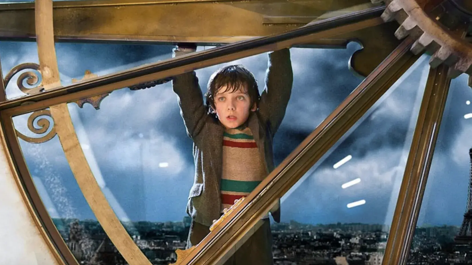 A young boy is hanging on the hand of a large clock on the outside of a building. 