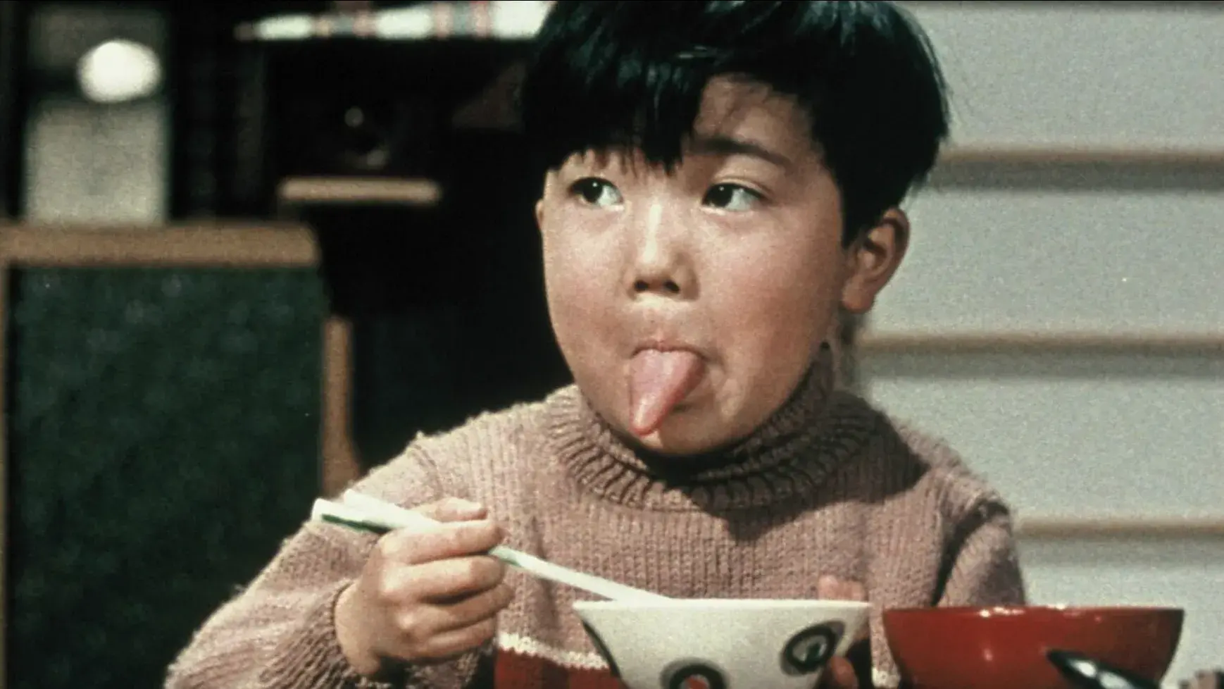 A young boy sits at a table holding chopsticks over a bowl with his tongue sticking out.