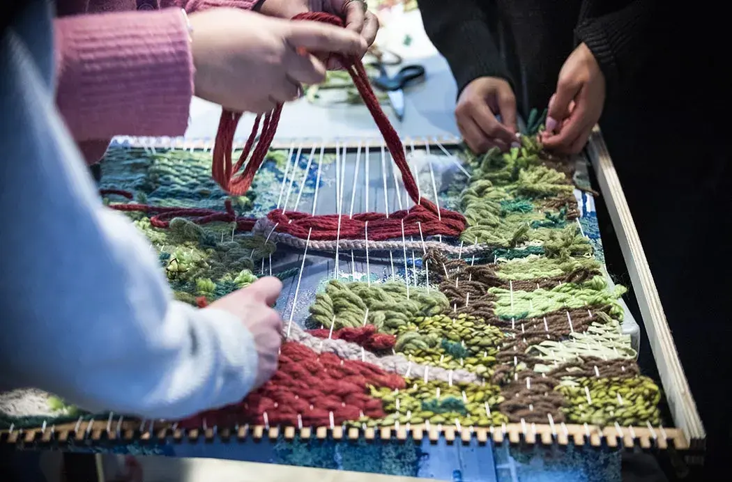 Hands weave thick yarn in various shades through a small tabletop loom