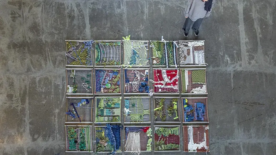 A person stands next to a grid of 20 weavings in small looms laid on a concrete floor