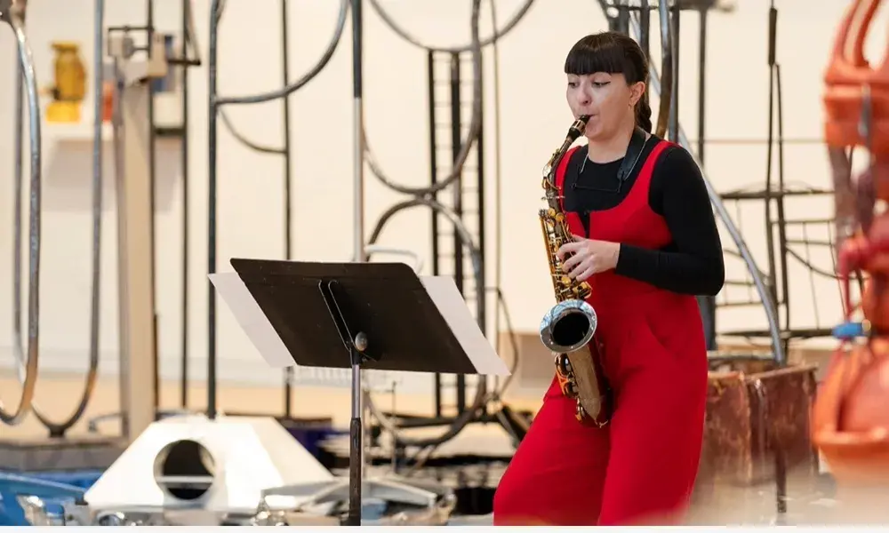 A woman wearing red overalls and a black shirt plays the saxophone with sculptures behind her