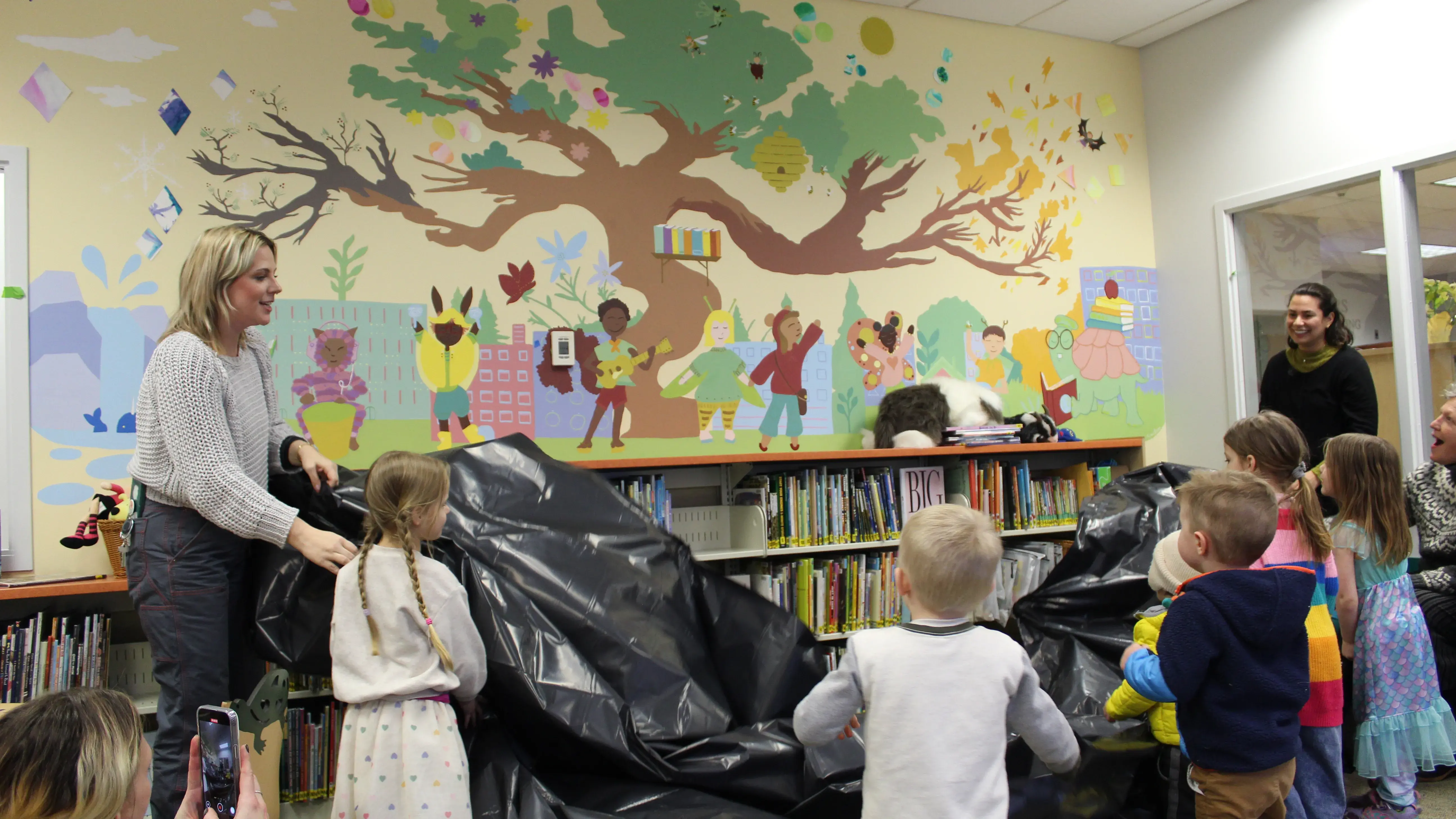 Pre-school children and their teachers remove a plastic covering to unveil a newly painted mural for the school library.