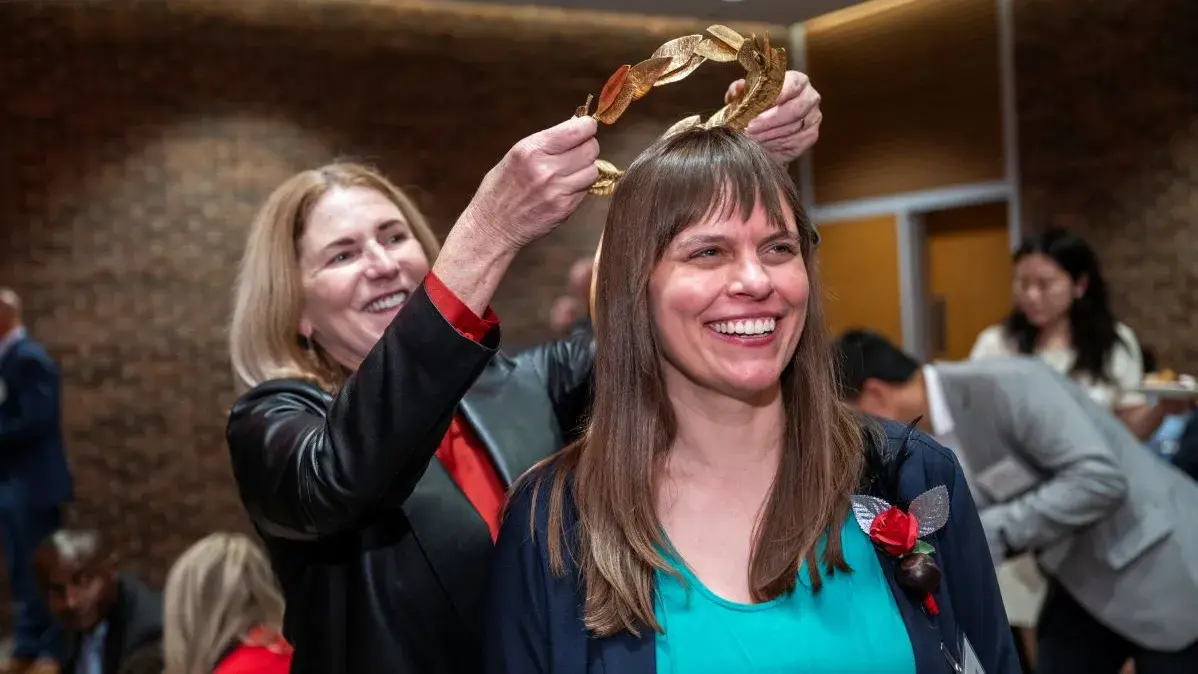 Kate Shannon, a white woman with long straight dark hair, stands while golden laurels are placed on her head
